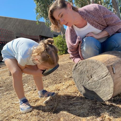 A preschool girl bends over to examine something on the ground with her magnifying glass. A CuriOdyssey staff member is crouched next to her on a log, explaining what she's seeing
