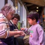 A young kid carefully pets a millipede held by a CuriOdyssey volunteer