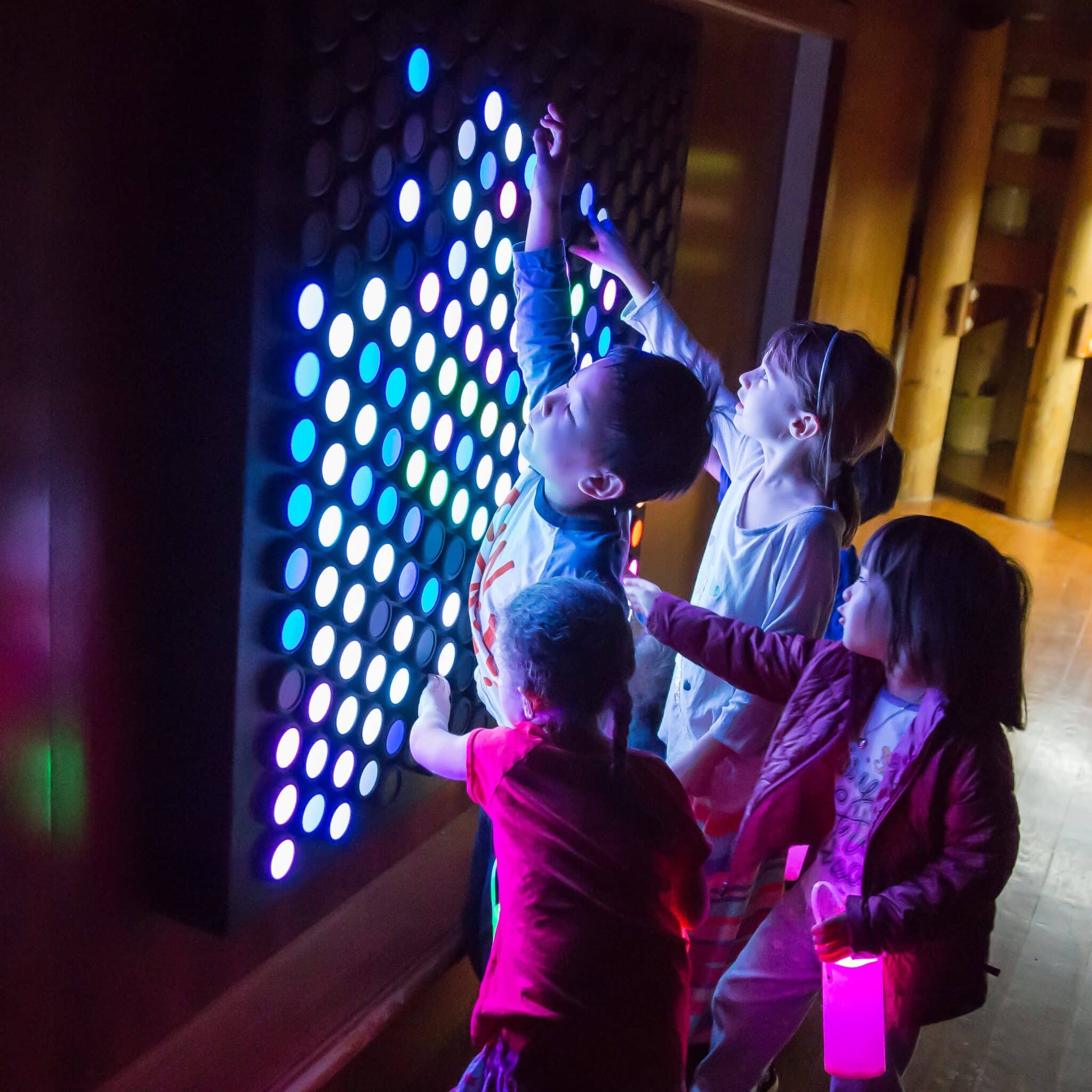 "Children joyfully interact with a vibrant multicolored light exhibit, their faces illuminated by the glowing hues. They reach out to touch the shifting colors, captivated by the dynamic patterns and playful atmosphere