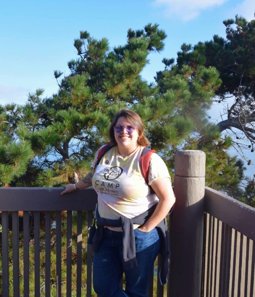 A photo of Emma, a person in a Camp CuriOdyssey t-shirt, jeans, and sunglasses. They have shoulder-length brown hair and are carrying a red backpack. They are standing in front of a railing with one hand on the railing and the other hand in their pocket. There are green pine trees in the background.