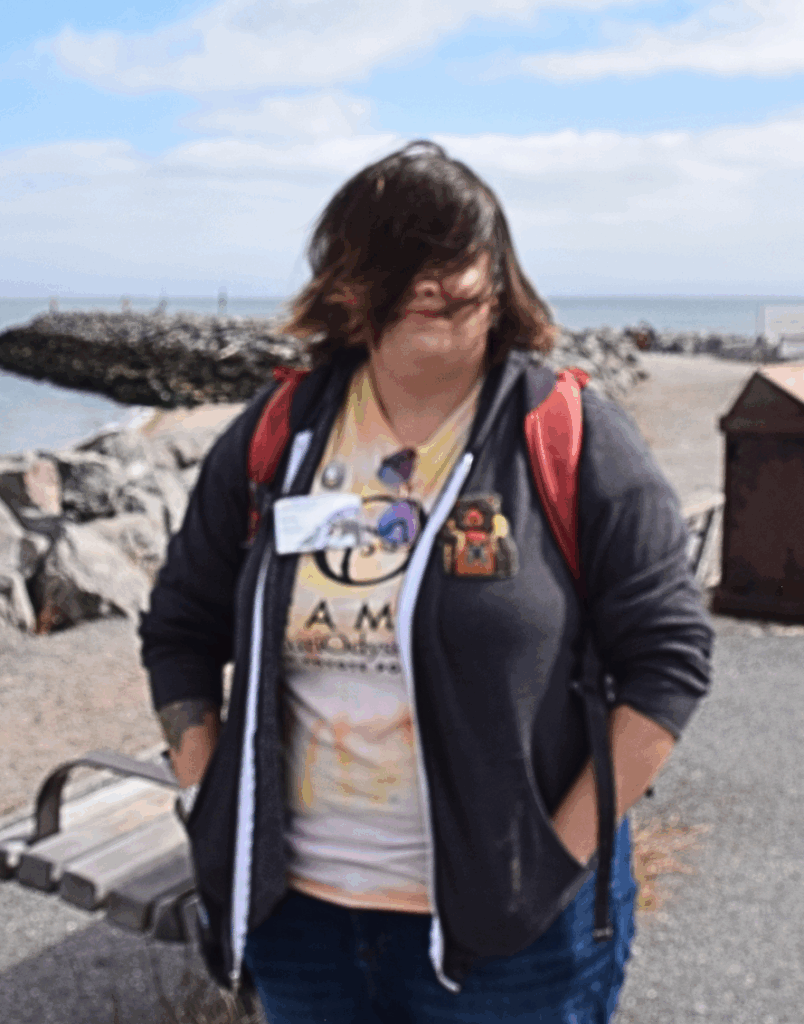  A photo of Emma, wearing a jacket, standing on the Jetty. Their hair is blowing all over their face. 
