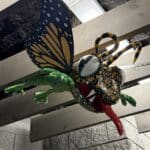 Up close image of the frog alebrije. It is multicolored, has monarch butterfly wings, and has its tongue out.